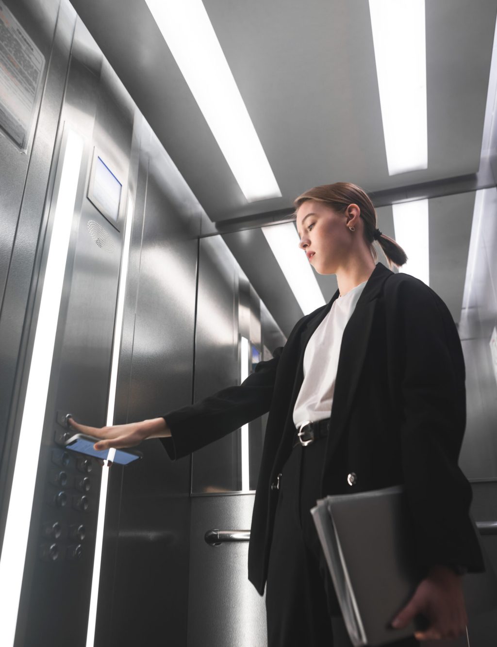Serious businesswoman pressing the button of the elevator holding smartphone in one hand and laptop in another. Female office worker is pushing the lift button and holding gadgets in her hands.